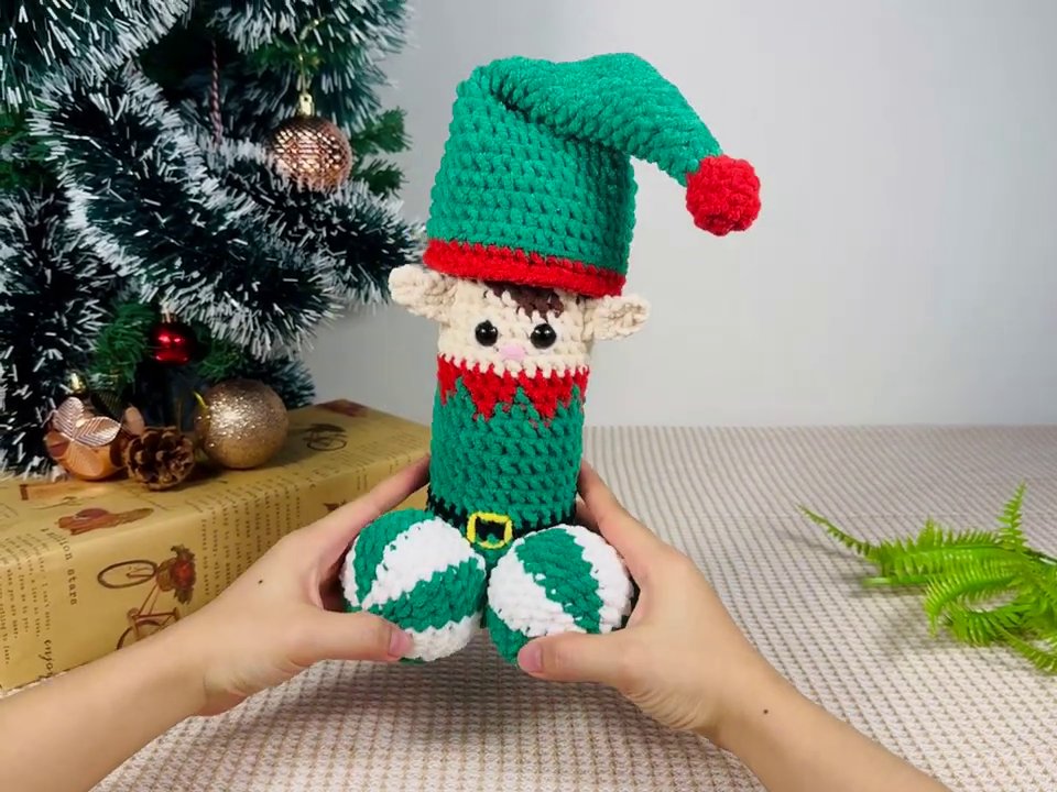 The Enchanting World of Handmade Crochet Elf Dolls: A Festive Showcase 2 The crochet elf doll is first presented, held between two hands against a backdrop of a decorated Christmas tree and a wrapped gift.